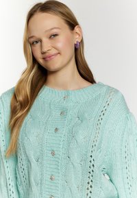 Maglione lavorato a maglia di colore azzurro chiaro con motivi a treccia, dotato di scollatura rotonda e bottoni decorativi sulla parte anteriore. Tessuto morbido e strutturato.