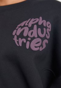 Čierna mikina s vyvýšeným fialovým textom "alpha industries." Tkanina má hladkú textúru, okrúhly výstrih a dlhé rukávy.