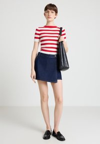 Frau mit kurzen braunen Haaren, die ein rot-weiß gestreiftes Hemd, einen dunkelblauen asymmetrischen Rock, schwarze Slipper trägt und eine schwarze Schultertasche bei sich hat.