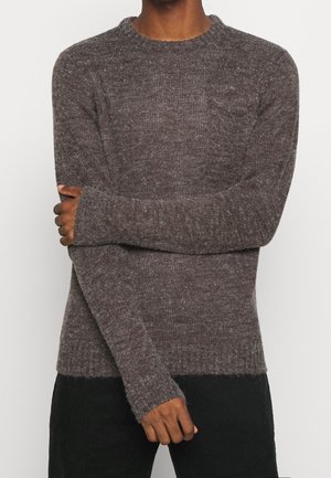 Personne aux bras croisés portant un pull en tricot marron et duveteux ainsi qu'un pantalon noir, sur un fond clair uni.