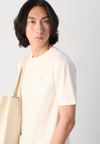 Cremfarbene Baumwoll-T-Shirt mit Rundhalsausschnitt und kurzen Ärmeln, mit einem kleinen türkisfarbenen Logo auf der oberen Brust. Tragetasche aus Canvas über der Schulter.