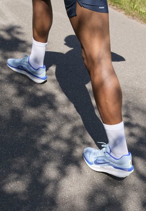 Des chaussures de course bleu clair présentent une tige en maille, des accents violets contrastants, une semelle blanche et une marque visible. Portées avec une chaussette blanche.