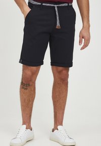 Navy blue shorts made of cotton blend with rolled cuffs, featuring a patterned belt and front pockets. Paired with white sneakers.