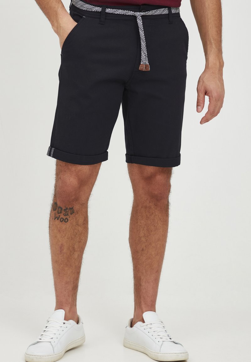 Navy blue shorts made of cotton blend with rolled cuffs, featuring a patterned belt and front pockets. Paired with white sneakers.