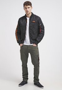 Jachetă bomber neagră cu accente portocalii, cu buzunare cu fermoar și manșete cu cusături, purtată cu pantaloni cargo verde măsliniu și adidași negri.