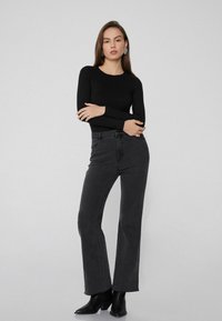Schwarzes Langarmoberteil kombiniert mit hochtragen schwarzen Jeans. Die Jeans haben einen geraden Schnitt und einen ausgefransten Saum; das Modell trägt schwarze Stiefeletten.
