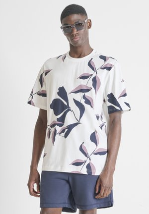 Mladić nosi pretjerano velike crne sunčane naočale, bijelu majicu s velikim ljubičastim i crnim uzorcima lišća te tamnoplave kratke hlače.