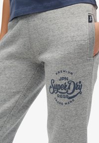 Pantalones de chándal grises con un acabado texturizado, que presentan un logo azul de "SuperDry" y una pequeña etiqueta de la marca. El diseño incluye un corte relajado y bolsillos laterales.
