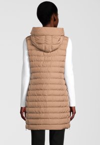 Chaleco acolchado beige con capucha, con textura de acolchado horizontal y cremalleras laterales. Lleva puesto un top de manga larga blanco, de diseño de longitud media.