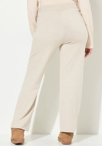 Vrouw in beige gebreide broek en bijpassende trui, staand met de rug naar de kijker, met bruin suède enkellaarsjes, op een witte achtergrond.
