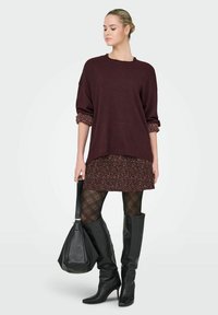 Przesadnie duży bordowy sweter z dzianiny nałożony na dopasowaną sukienkę w kwiaty, połączony z czarnymi rajstopami w kratę oraz czarnymi skórzanymi kozakami do kolan.