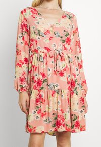 Žena v růžových květinových šatech s dlouhými rukávy, páskem v pase a výstřihem do V, zobrazená od ramen po střed stehna.