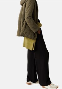 Olivově zelená prošívaná bunda s kapucí, nošená přes žlutý žebrovaný svetr, doplněná širokými černými kalhotami a krémovými teniskami s oranžovými akcenty.