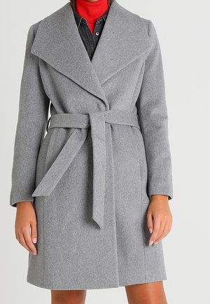 Kvinde iført en knælængde, grå uldfrakke med bælte over en rød højhalset trøje og en sort skjorte, stående med hænderne ned langs siden.