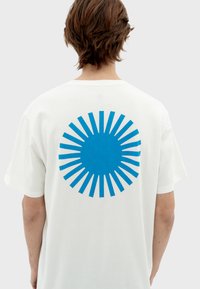 Witte katoenen t-shirt met een groot blauw cirkelvormig ontwerp en stralende accenten op de achterkant. Korte mouwen en ronde halslijn.