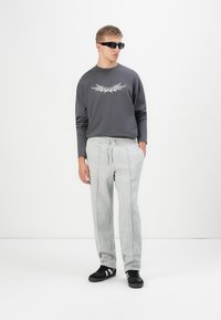 Joven con gafas de sol negras, camiseta de manga larga gris oscuro con un gráfico, pantalones de chándal gris claro y zapatillas negras, de pie contra un fondo blanco.