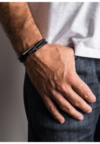 Brăță din piele împletită neagră cu un închizător din metal argintiu. Închizătoarea are inscripții gravate și are o formă stilizată, dreptunghiulară.