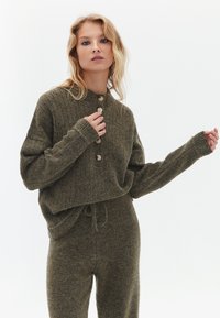 Olivgrüner Strickpullover mit gerippter Textur, Knopfverschluss und langen Ärmeln, kombiniert mit passenden Jogginghosen.