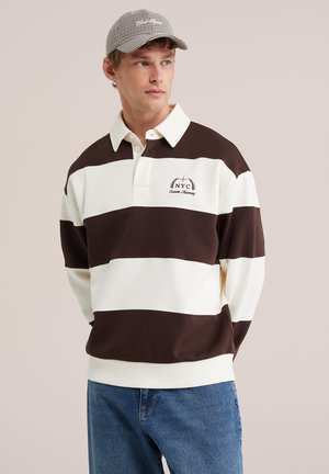 Jeune homme portant un maillot de rugby rayé marron et blanc avec broderie NYC et une casquette à carreaux, debout les mains dans le dos.