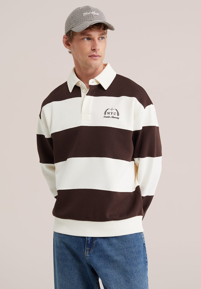 Jeune homme portant un maillot de rugby rayé marron et blanc avec broderie NYC et une casquette à carreaux, debout les mains dans le dos.