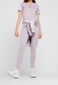 Lila idrottsoutfit med en åtsittande T-shirt och leggings, med vita ränder på ärmarna och en knuten sweatshirt i midjan.