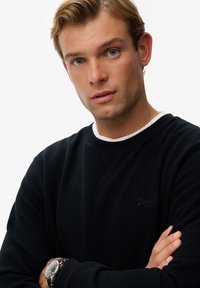 Sort sweatshirt med ribbet krave og manchetter, med et lille logo på brystet. Model bærer et sølvur, med armene foldet.