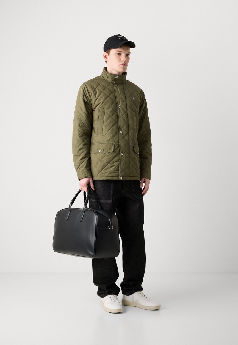 Olive drab pikowana kurtka z zatrzaskami, dwie przednie kieszenie, czarna skórzana torba, czarne dżinsy i białe trampki. Czarna czapka z logo.