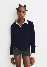 Maglione a maglia blu navy con una camicia a righe bianche e grigie sotto, abbinato a pantaloni corti grigi e una cintura nera con doppia fibbia.