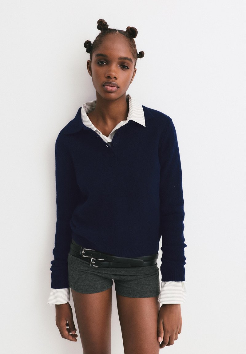 Maglione a maglia blu navy con una camicia a righe bianche e grigie sotto, abbinato a pantaloni corti grigi e una cintura nera con doppia fibbia.
