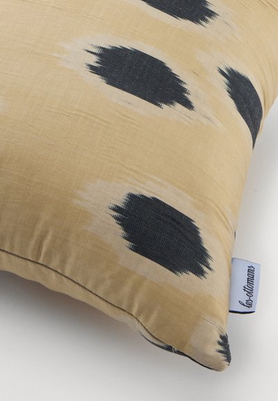Coussin carré beige avec des motifs irréguliers de coups de pinceau noirs et une étiquette en tissu blanc sur le côté avec un texte foncé.