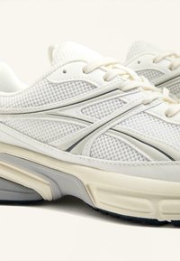 Bijele atletske cipele s mrežastim gornjim dijelom, glatkim kožnim akcentima, zaobljenim prstima i teksturiranim đonom s crnim gumiranim gaznim slojem.