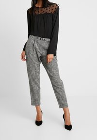 Czarna bluzka z koronkowymi detalami przy dekolcie, w połączeniu z szarymi teksturowanymi spodniami. Czółenka na wysokim obcasie w kolorze czarnym dodają kontrastu.