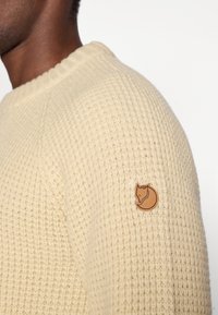 Pull en tricot crème avec un motif texturé et un col côtelé, doté d'un patch circulaire de logo marron sur l'épaule gauche.