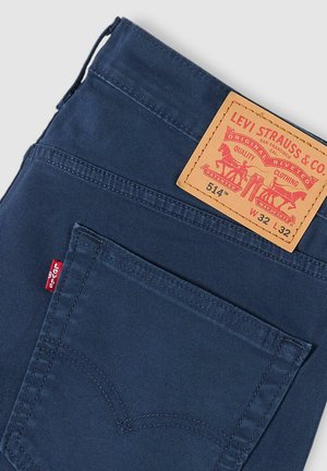 Pantalones de mezclilla azul con un parche trasero de cuero marrón etiquetado "Levi Strauss & Co.", con costuras rojas y una pequeña etiqueta roja en el bolsillo.