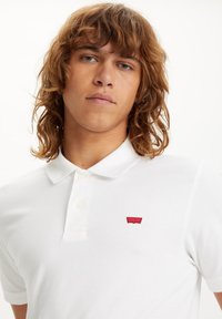 White polo shirt made of cotton, featuring a two-button placket and a small red logo on the chest. Smooth texture and classic fit.