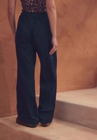 Navybukser med vide ben, elastik i taljen og baglommer, kombineret med mørkt fodtøj, mod en terracottafarvet baggrund.