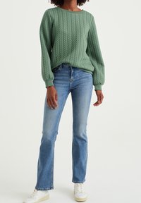 Grüner, strukturiert gestrickter Pullover mit gesteppter Musterung, rundem Ausschnitt und langen Ärmeln, kombiniert mit blauen Schlagjeans und weißen Turnschuhen.