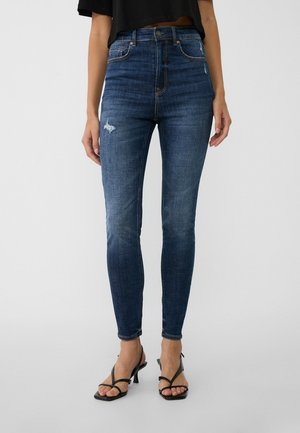 Femme portant un jean skinny taille haute bleu foncé légèrement déchiré et des sandales noires à talons avec brides sur un fond uni.