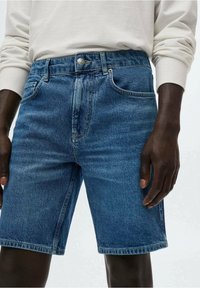 Shorts en denim bleu moyen avec une coupe classique. Comprend un bouton à l'avant, des poches traditionnelles et des coutures visibles le long des coutures.