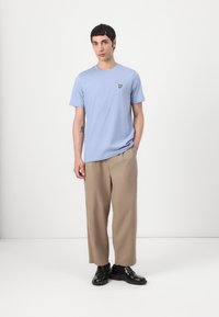 T-shirt bleu clair à manches courtes avec un logo brodé jaune sur la poitrine, associé à un pantalon large beige et des chaussures noires.