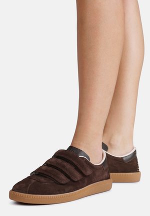 Adidași maro din piele întoarsă cu trei benzi Velcro și talpă din cauciuc, purtați pe picioare goale, văzuți din unghi lateral și din spate.