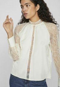 Blusa de marfil con mangas largas de encaje transparentes y un cuello alto. El cuerpo es liso, con un frente abotonado y delicados detalles de encaje.