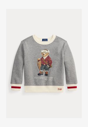 Harmaa huppari, jossa on karhu punaisessa flanellipaidassa, joka pitää kädessään mäkiä. Resorit vaaleassa kermassa punaisilla yksityiskohdilla, logotagi helmaosassa.