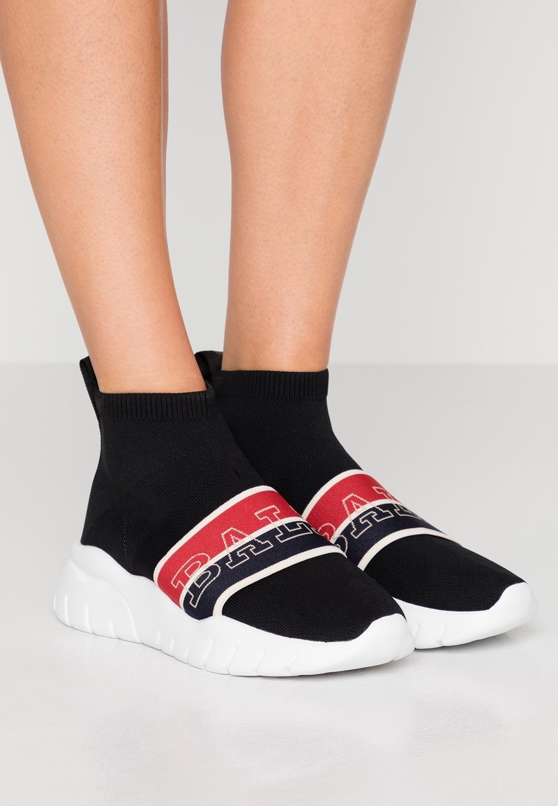 Jambes portant des baskets noires style chaussettes avec semelles blanches et bandes rouges, blanches et bleues avec un texte en gras sur le dessus.