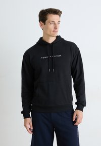 Mann iført en svart Tommy Hilfiger hettegenser og marineblå joggebukser, står mot en enkel hvit bakgrunn og ser til siden.