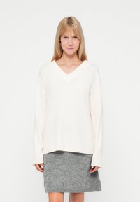 Krémový sveter s výstrihom do V a dlhými rukávmi, s rebrovanými manžetami a lemom, skombinovaný s textúrovanou sivou sukňou. Mäkký pletený materiál, voľný strih.