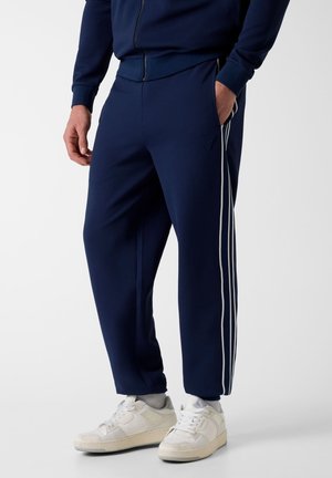 Homme portant un pantalon de survêtement bleu marine texturé avec des bandes blanches sur les côtés, des chaussettes blanches, des baskets blanches et une veste zippée assortie bleu marine.
