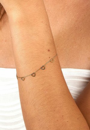 Delikat guldkædearmbånd med fire små åbne hjertecharms, båret på en underarm med lysebrun hud og en hvid tekstureret top.