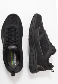 Zapatillas deportivas negras que presentan materiales de malla y sintéticos, con una plantilla acolchada, suela texturizada y una lengüeta en el talón.