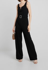 Mono negro con escote en V profundo, piernas anchas y un cinturón en la cintura con una hebilla cuadrada. Modelo sosteniendo un pequeño bolso con patrón de piel de serpiente.